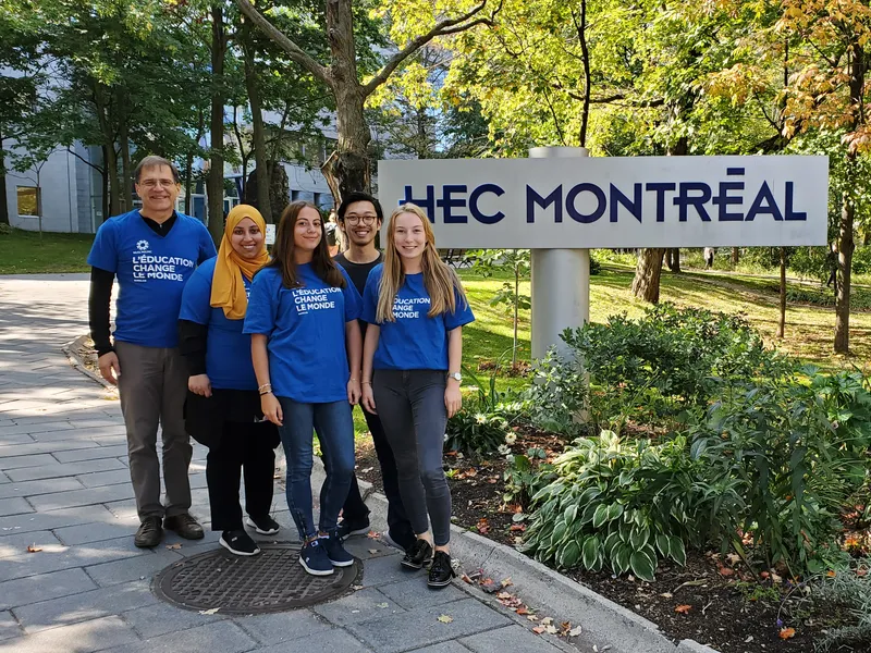 Five members of the PÉR HEC committee in WUSC T-shirts beside the HEC Montréal sign